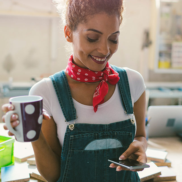 Young woman holding a cup of coffee while looking at her smartphone in her workshop