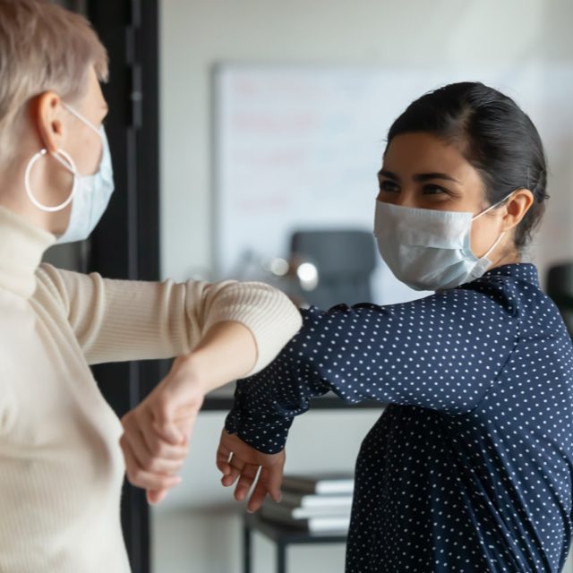 Duas mulheres utilizando máscaras de proteção sobre nariz e boca, cumprimentando-se com toque de cotovelo