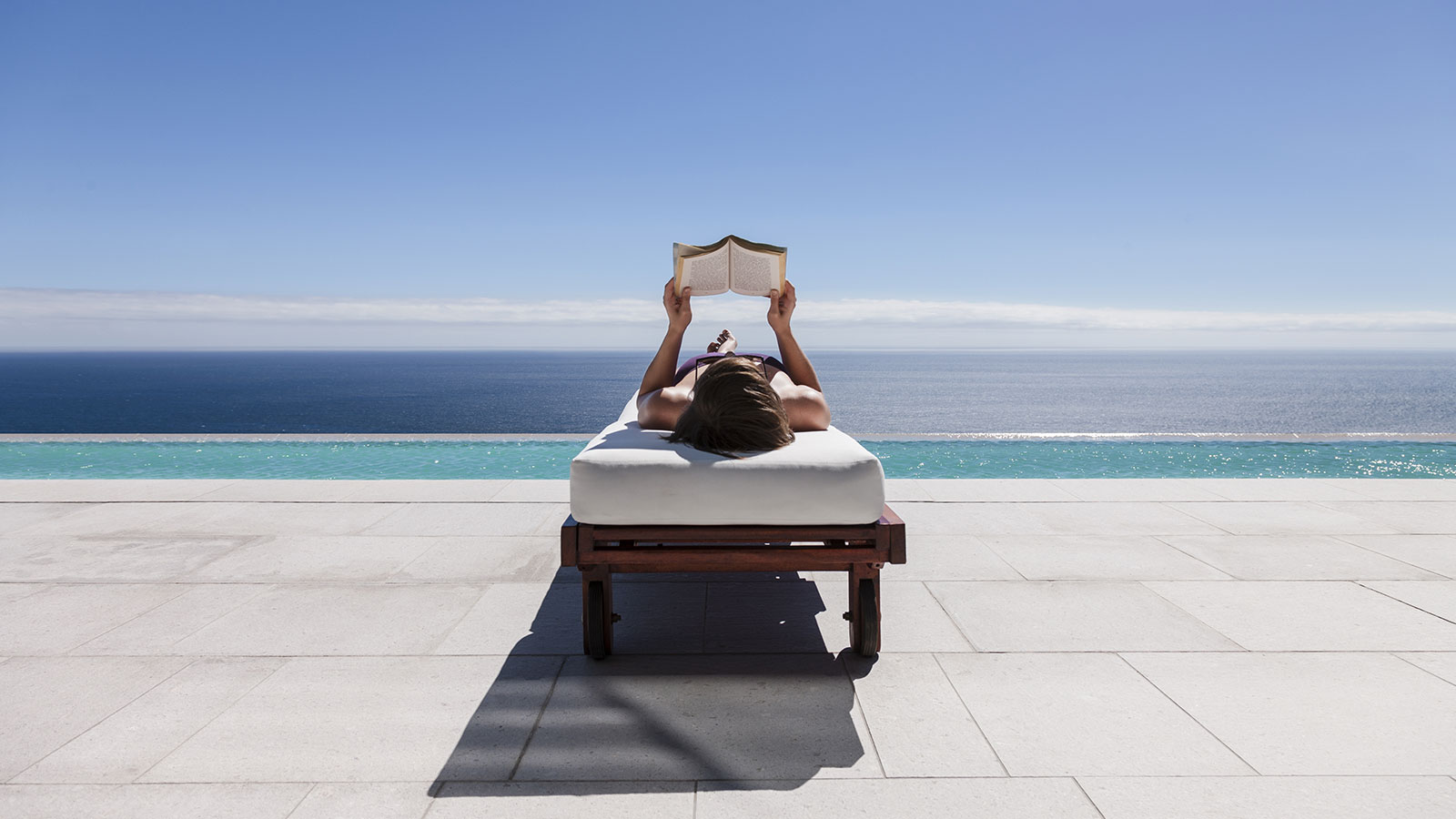 A woman reading a book while reclining on a lounge chair overlooking the beach.